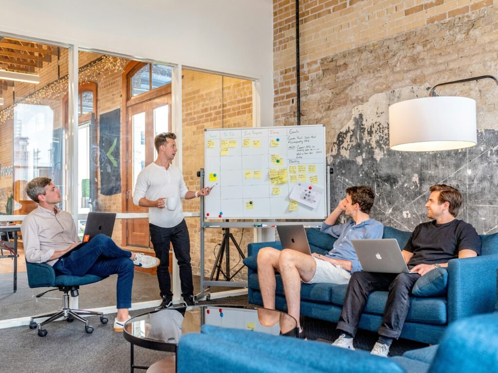 A modern office with a cozy, inviting atmosphere. A man stands in front of a whiteboard filled with colorful sticky notes, discussing something with three colleagues seated on a blue couch. The man in the center, wearing a blue shirt and white shorts, leans back with his hand on his head, while the man next to him, in a black shirt, listens attentively with his laptop open. Another man, in a light pink shirt, sits with his legs crossed, holding a laptop. The room features a brick wall, large glass doors, and a stylish glass table with a round base, reflecting the scene below.