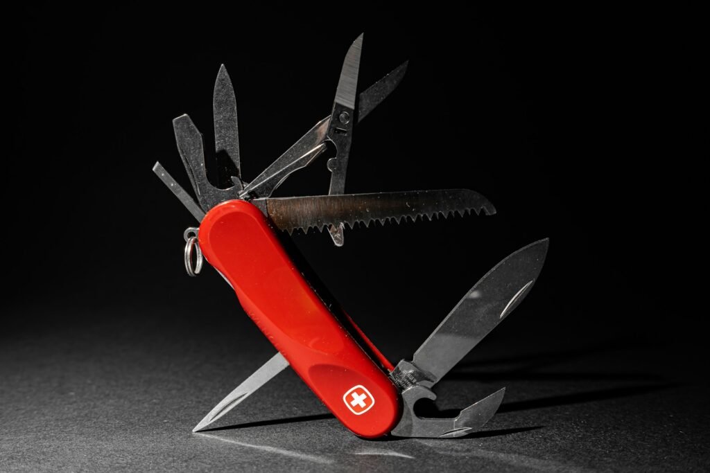 Multi-tool with red handle and various metal blades. The tool has a saw, several knives, and other implements. Swiss Army-style multi-tool with a red plastic handle and multiple metal blades, including a saw, displayed on a dark surface. A small ring is visible on the handle.