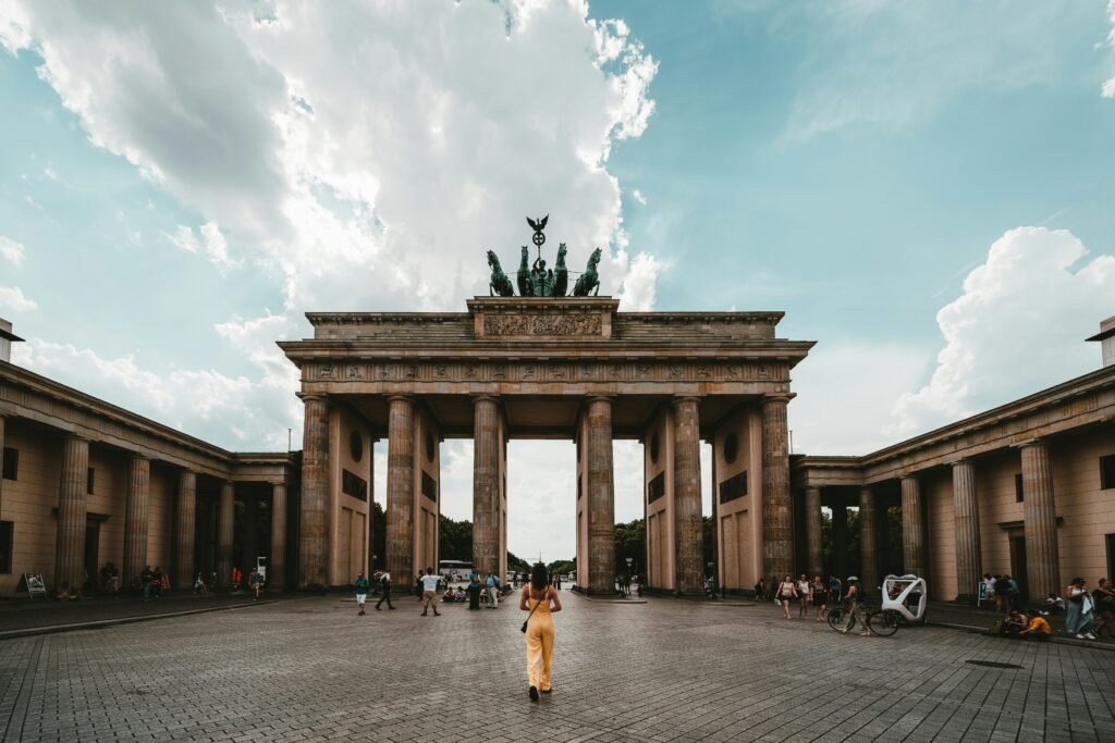 Brandenburg Gate in Berlin, with large, light-brown columns and an archway. People walk around the monument. A woman in a yellow jumpsuit is visible in the center of the image; a cloudy sky is above the gate.