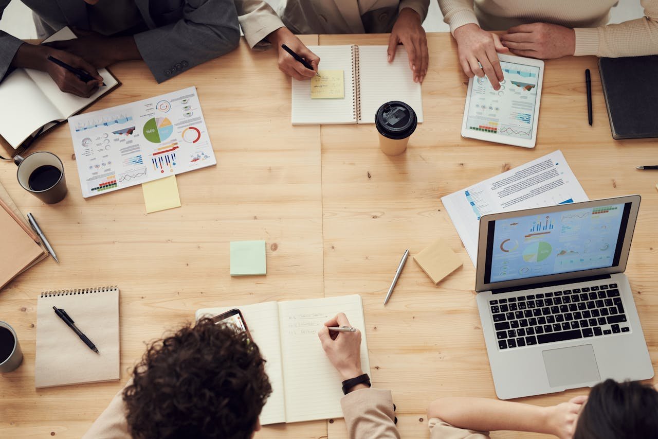 Diverse content team collaborating around a conference table with laptops, notebooks, and digital displays showing analytics dashboards in modern office setting