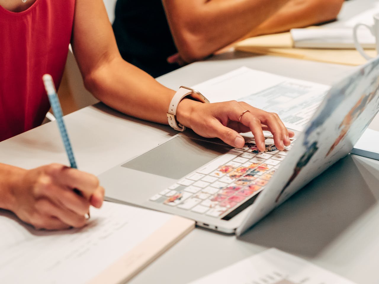Person in red shirt typing on laptop with colorful stickers while holding blue pen