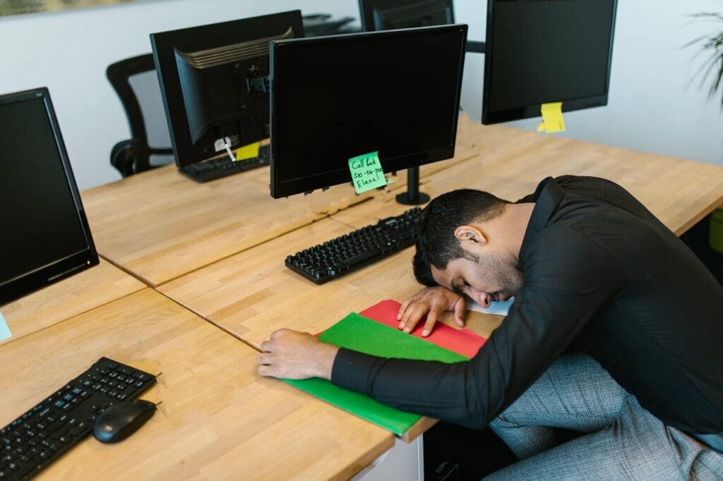 Man asleep at a wooden desk, head resting on the surface. Multiple computer monitors and keyboards are visible. A sticky note reads "Call back 580-10-386 Elena". He wears a black shirt and gray pants.