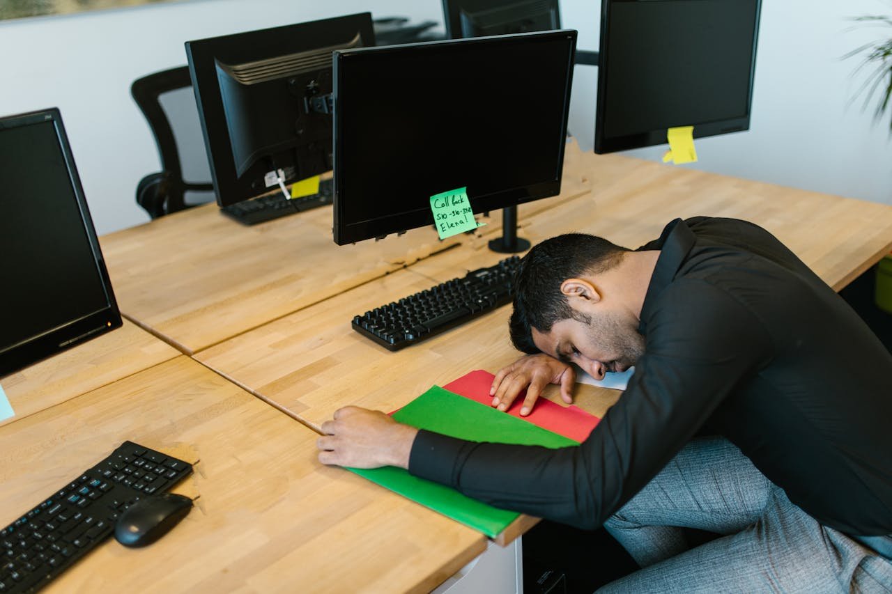 Man in black shirt lying head down on desk next to computer monitors with sticky note