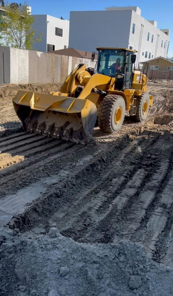 Yellow construction excavator working on a dirt excavation site, with a pile of dark gray dirt and parallel tire tracks. Modern light-colored buildings are in the background.