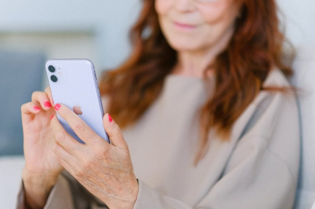 A person holds a light purple smartphone. Pink fingernails are visible. The person's upper body and hair are also in the image, but out of focus.