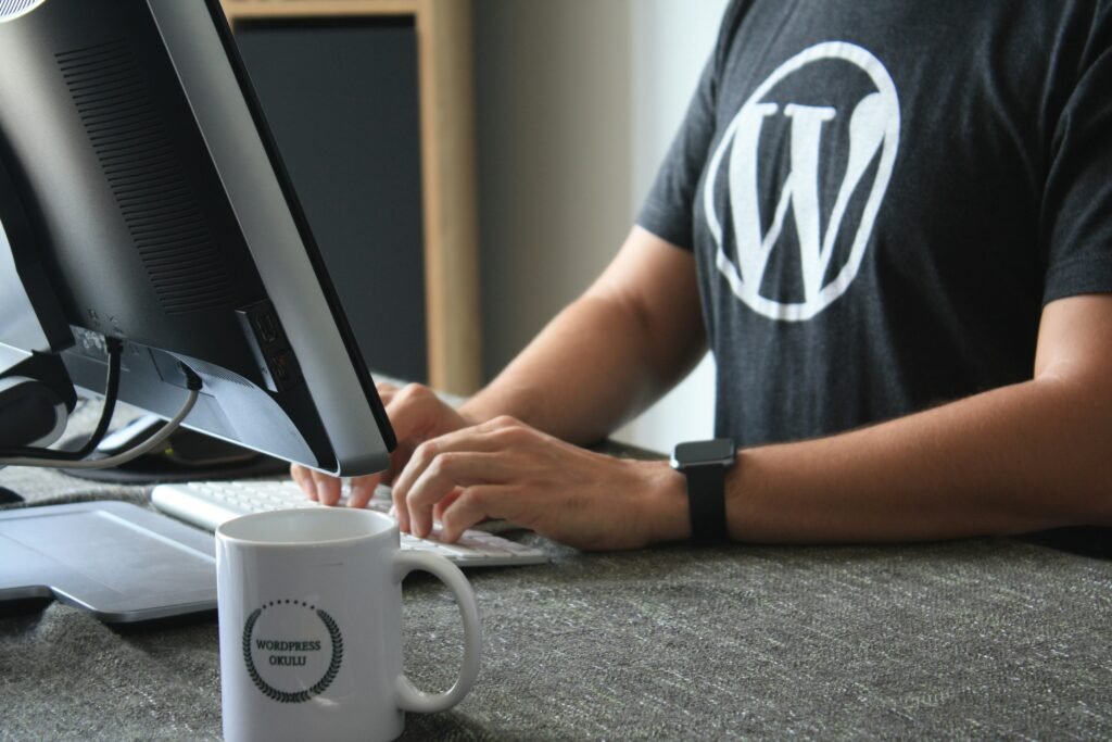 A person sits at a desk, typing on a white keyboard, with their right hand on the spacebar. They wear a dark grey t-shirt featuring a white 'W' logo. Beside them, a coffee mug reads "WordPress Okul" in black text. Their fingers are positioned over the keys, and a monitor stands behind them, partially visible. The desk surface is a muted grey, and a small portion of a wooden cabinet is visible in the background.