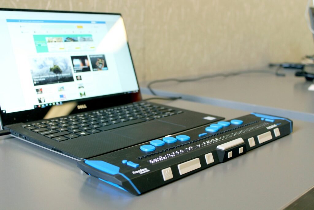 A sleek black laptop with a silver Dell logo rests on a polished gray desk. Its screen displays a webpage with various colorful images and articles. Adjacent to the laptop is a tactile device, primarily black and blue, with a series of large, textured buttons that are predominantly blue. The device, likely a specialized input tool, has a smooth, ergonomic shape and is positioned close to the laptop's keyboard. A beige wall with a subtle pattern provides a muted backdrop.